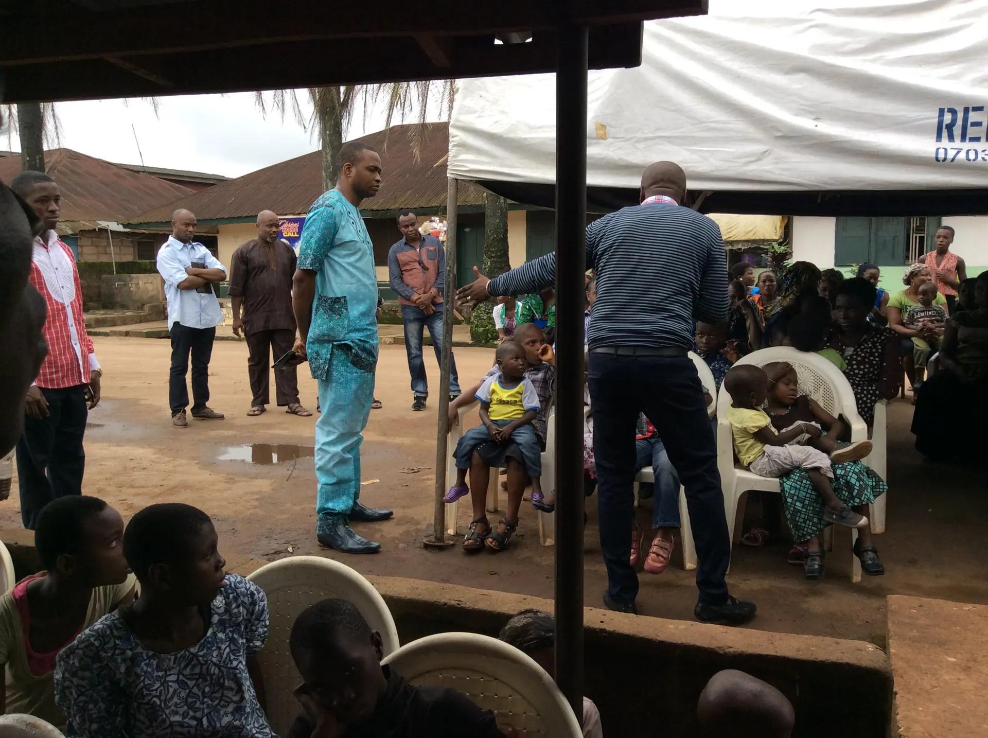 Community members and children seated under a white canopy while outreach volunteers coordinate the day's activities.