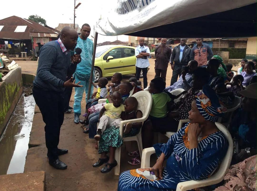 A foundation representative speaks with seated mothers and children at an open-air community gathering.