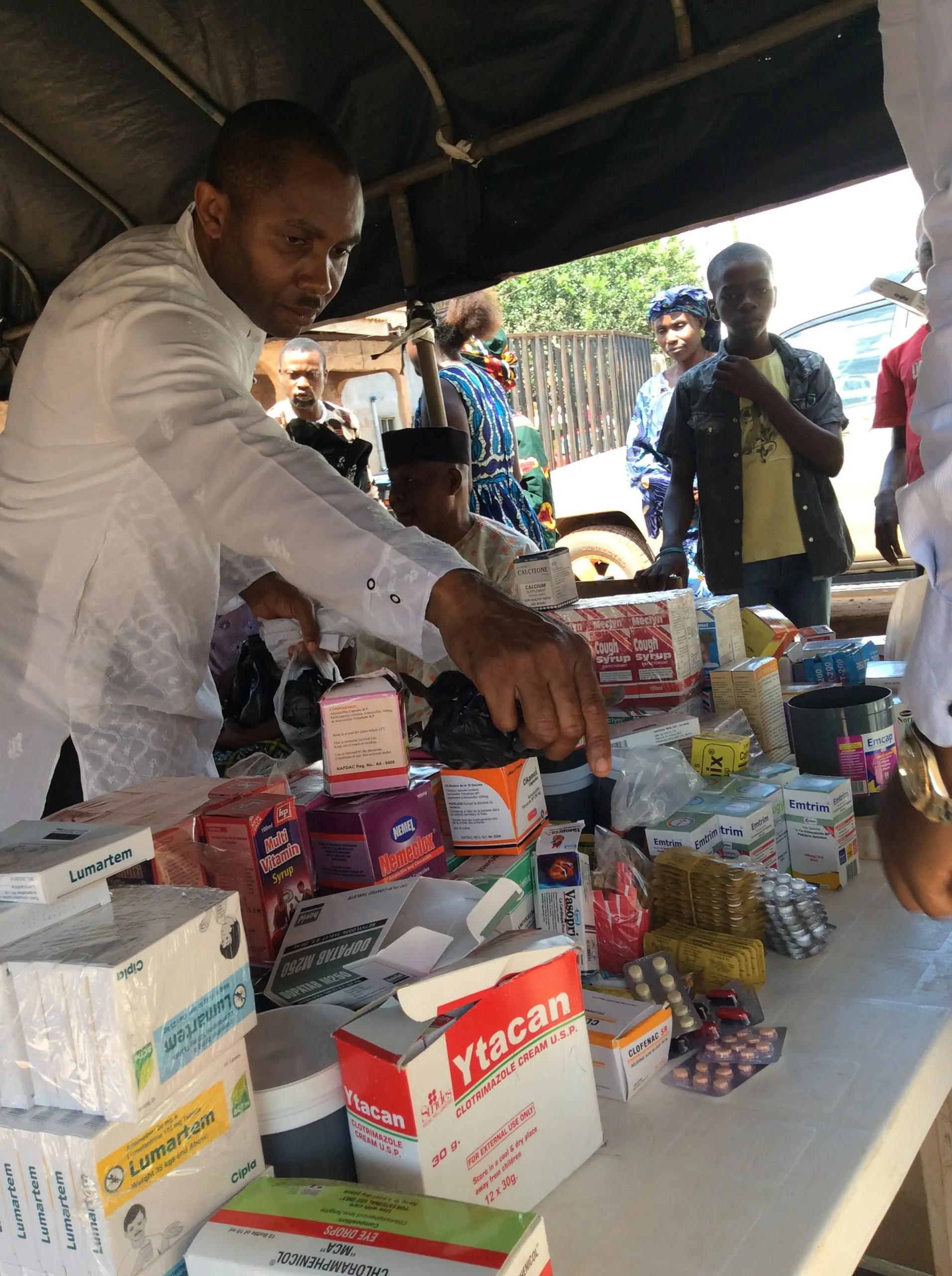 A volunteer in white sorts through boxes of medicines under a tent at a community outreach.