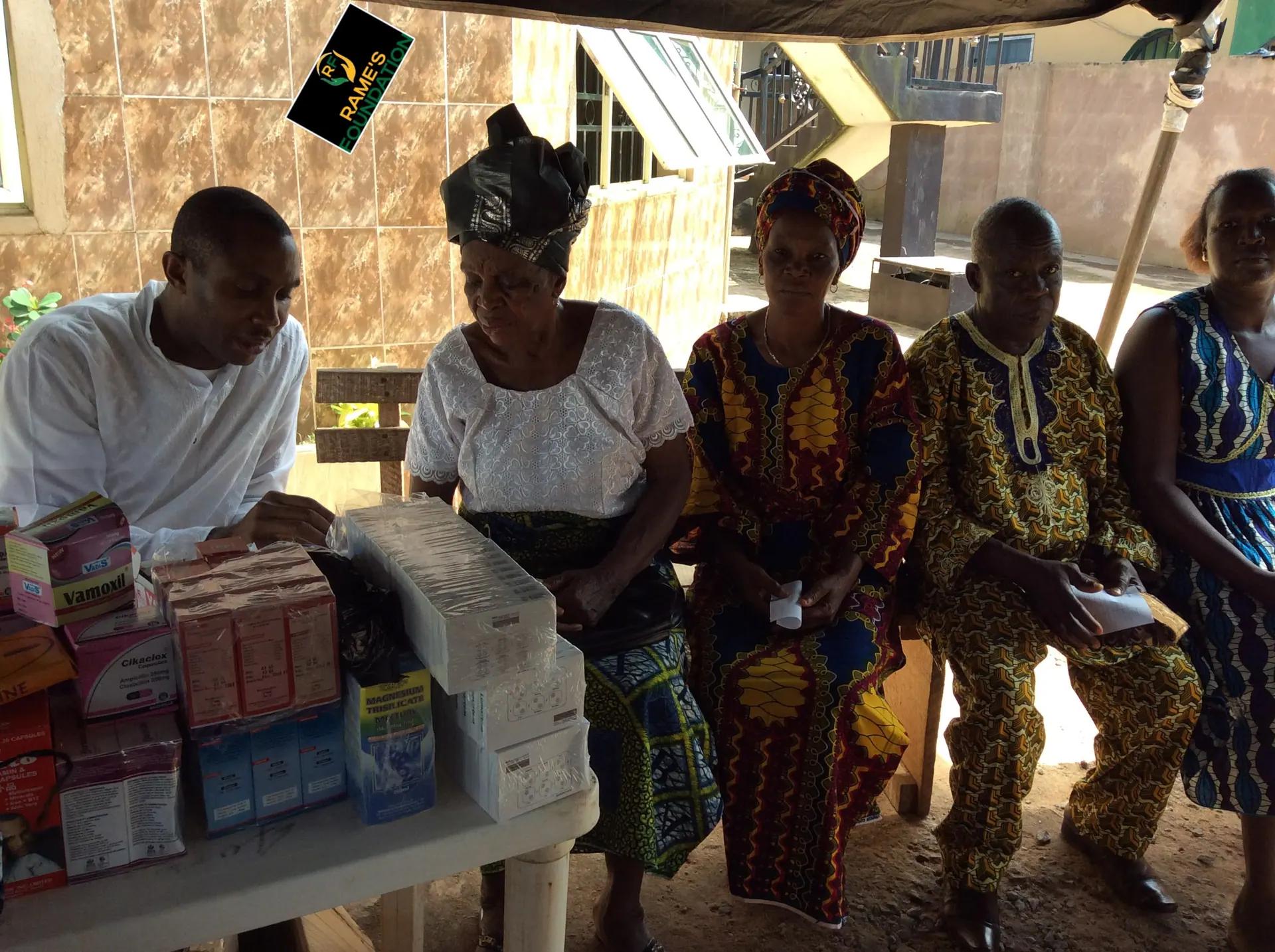 A volunteer at an outdoor table of medicines attends to seated women at a community outreach.