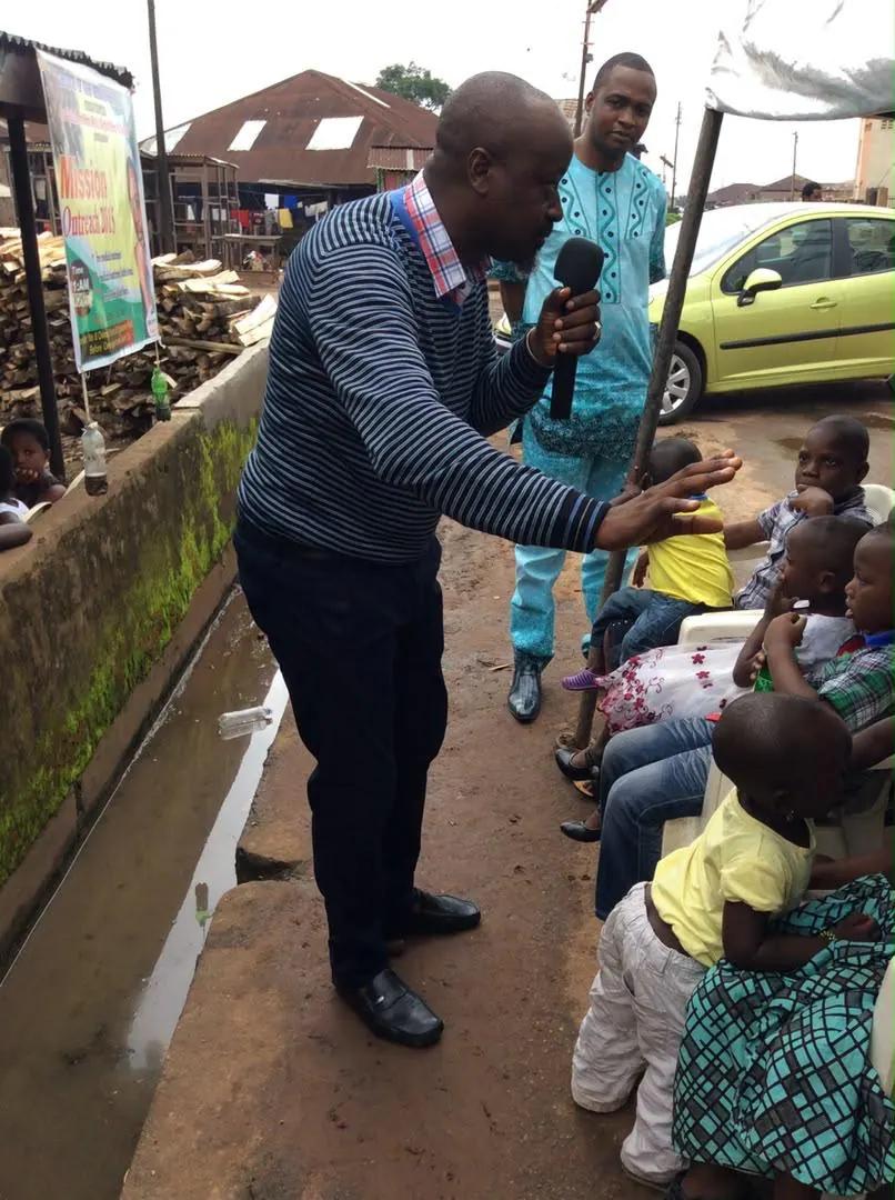 A RAMES team member with a microphone leans towards seated children at an outdoor outreach.