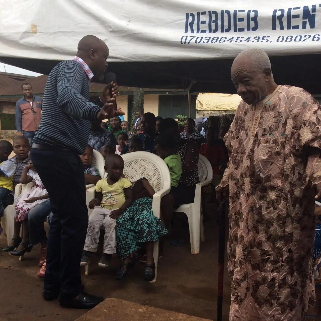Community members of all ages seated under a canopy listen to a RAMES team member.