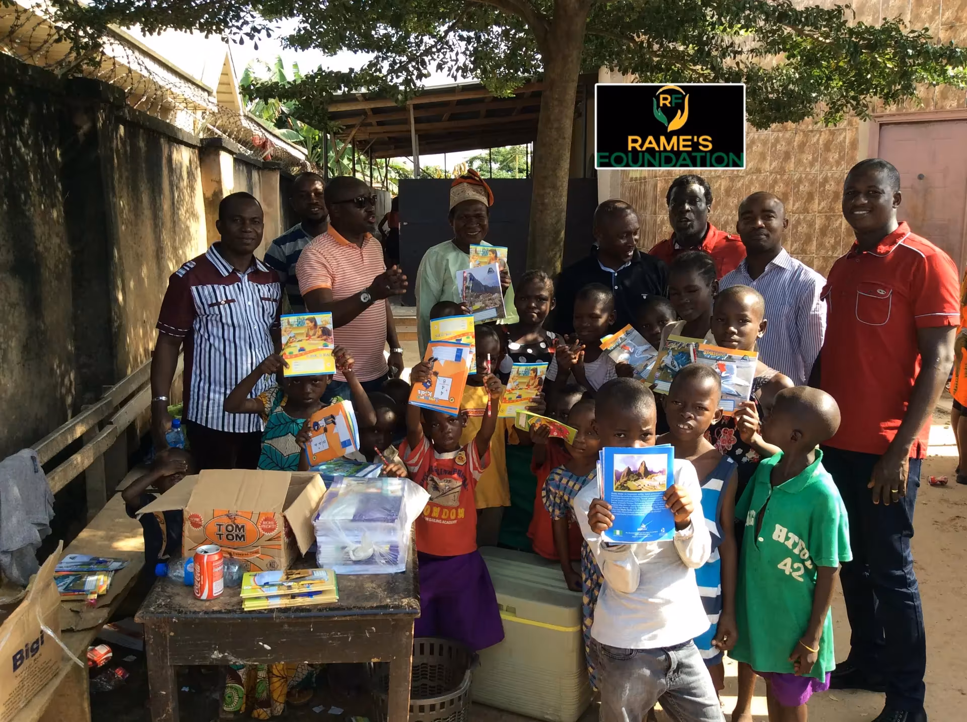 A group of smiling children raise their new exercise books beside RAMES Empowerment Foundation volunteers.