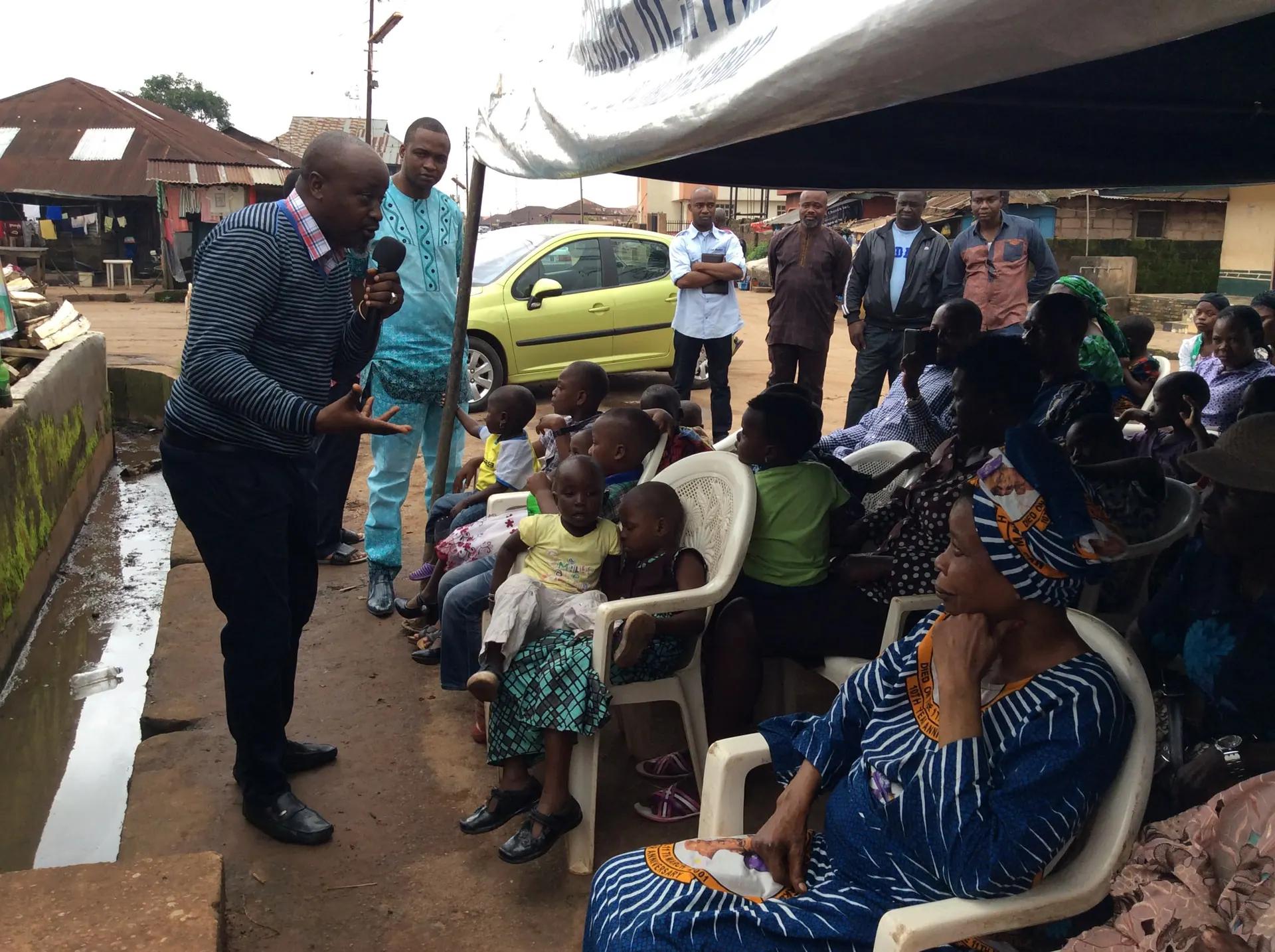 A RAMES Empowerment Foundation team member speaks into a microphone to families and children seated under a canopy at a community outreach in Lagos.