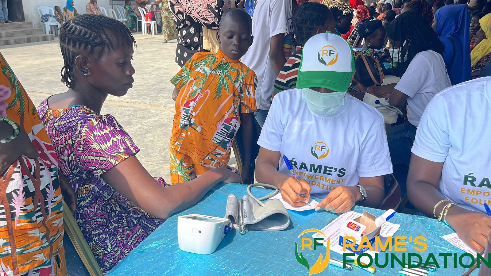 A masked volunteer in a RAMES cap checks a woman's blood pressure while other residents wait their turn.