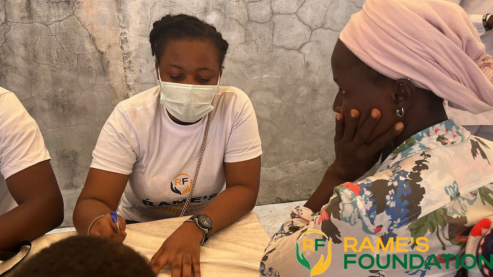 A masked volunteer listens to an elderly woman in a headscarf at a consultation table.