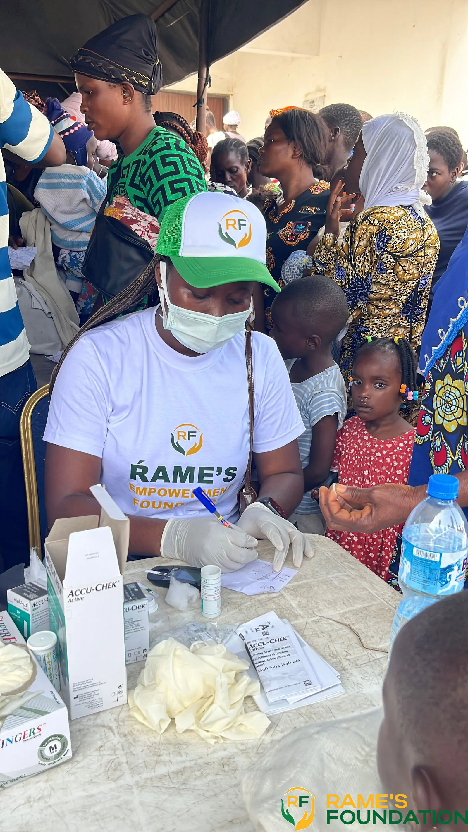 A masked RAMES volunteer works at the medicine dispensing table with residents gathered behind.