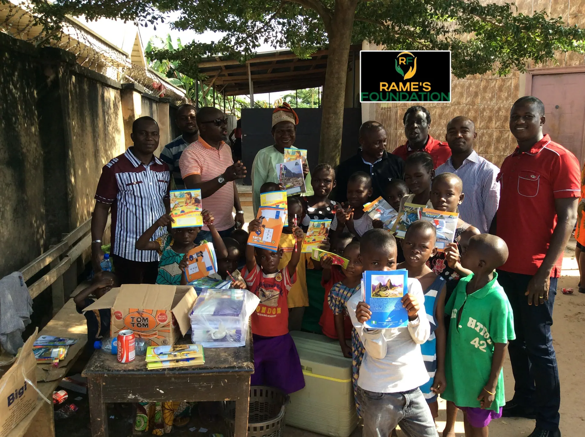 A group of smiling children raise their new exercise books beside RAMES Empowerment Foundation volunteers.