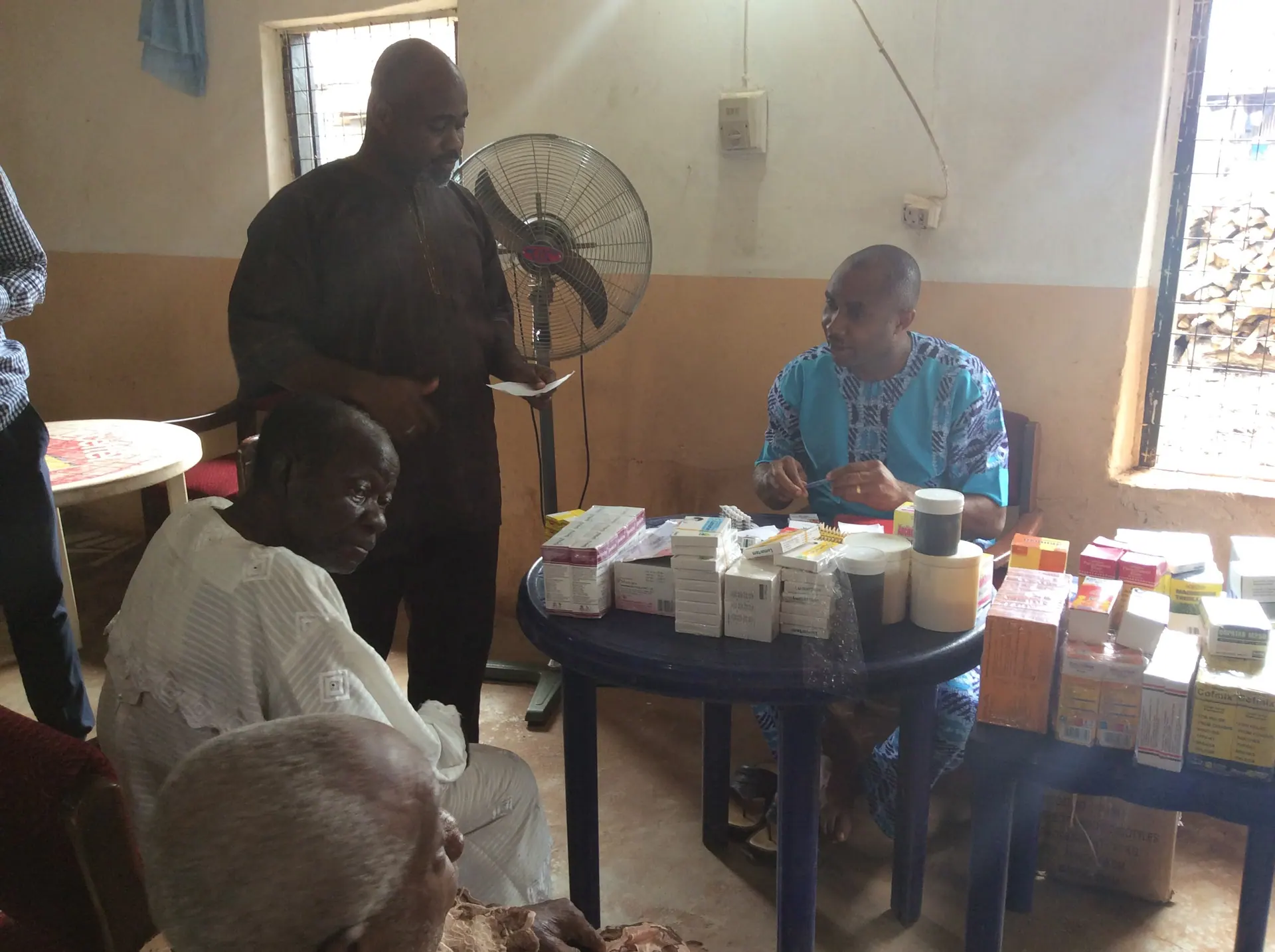 Volunteers sort medicines at a table while elderly community members wait to be attended to.
