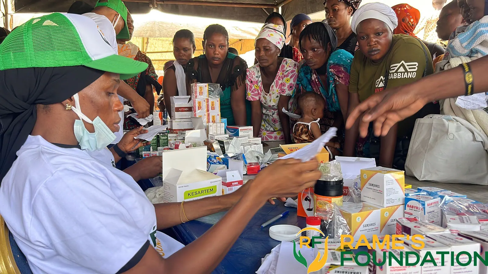 A masked volunteer reads a medicine pack at a table stacked with boxes of medicines while residents look on.