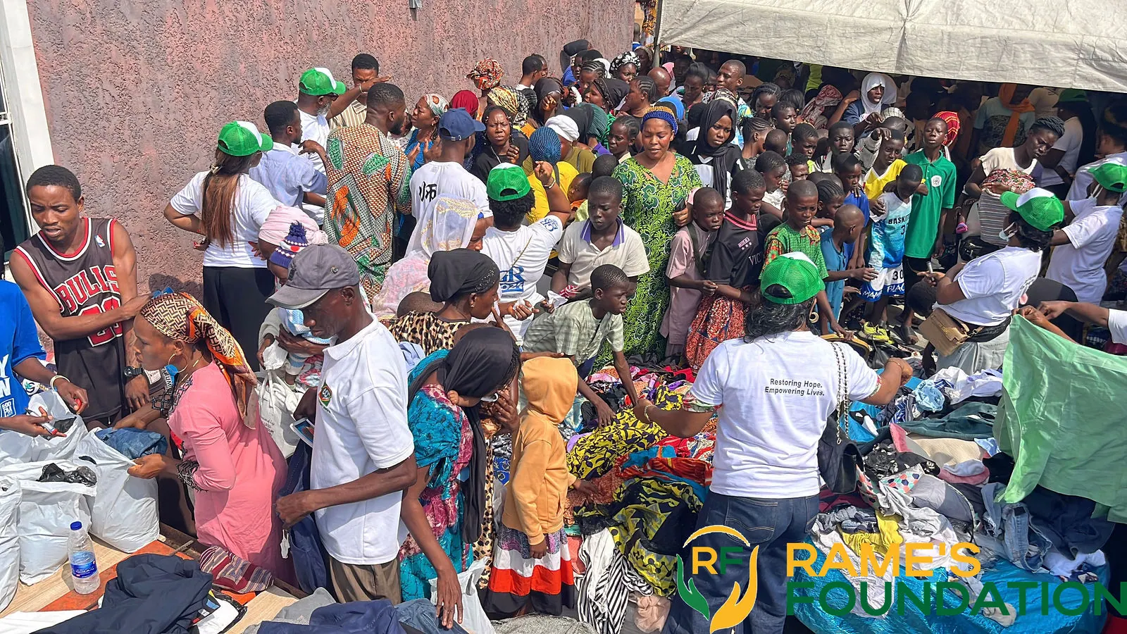A large crowd of residents gathers around tables of donated clothing under white canopies while RAMES volunteers in green caps hand out items.