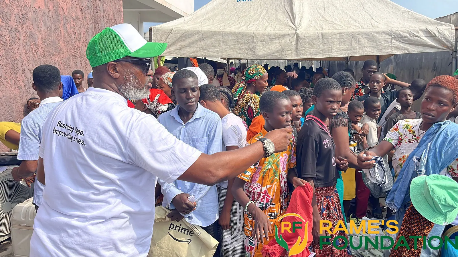 A volunteer in a RAMES cap reaches out to a group of children under a white tent.