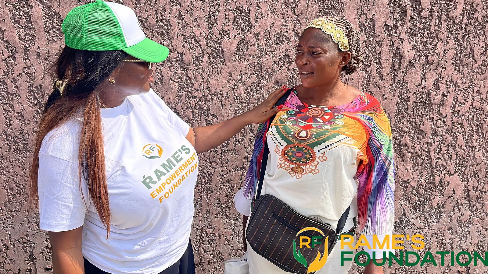 A RAMES volunteer in a green cap places a reassuring hand on the shoulder of an elderly woman in a patterned outfit.